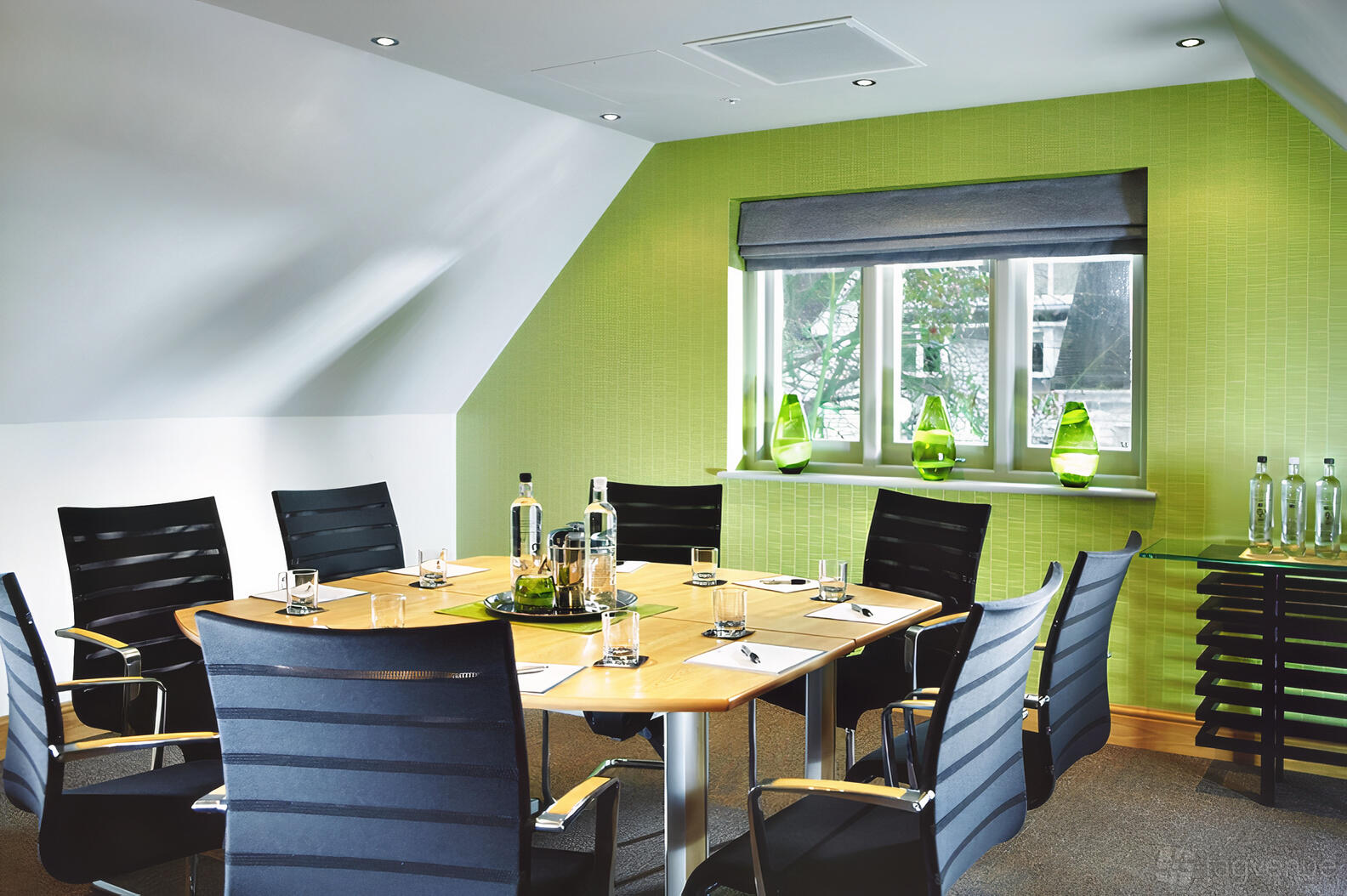 A hotel meeting room with a round wooden conference table, black chairs, green accent wall, and natural light at Rowhill Grange Hotel & Utopia Spa.