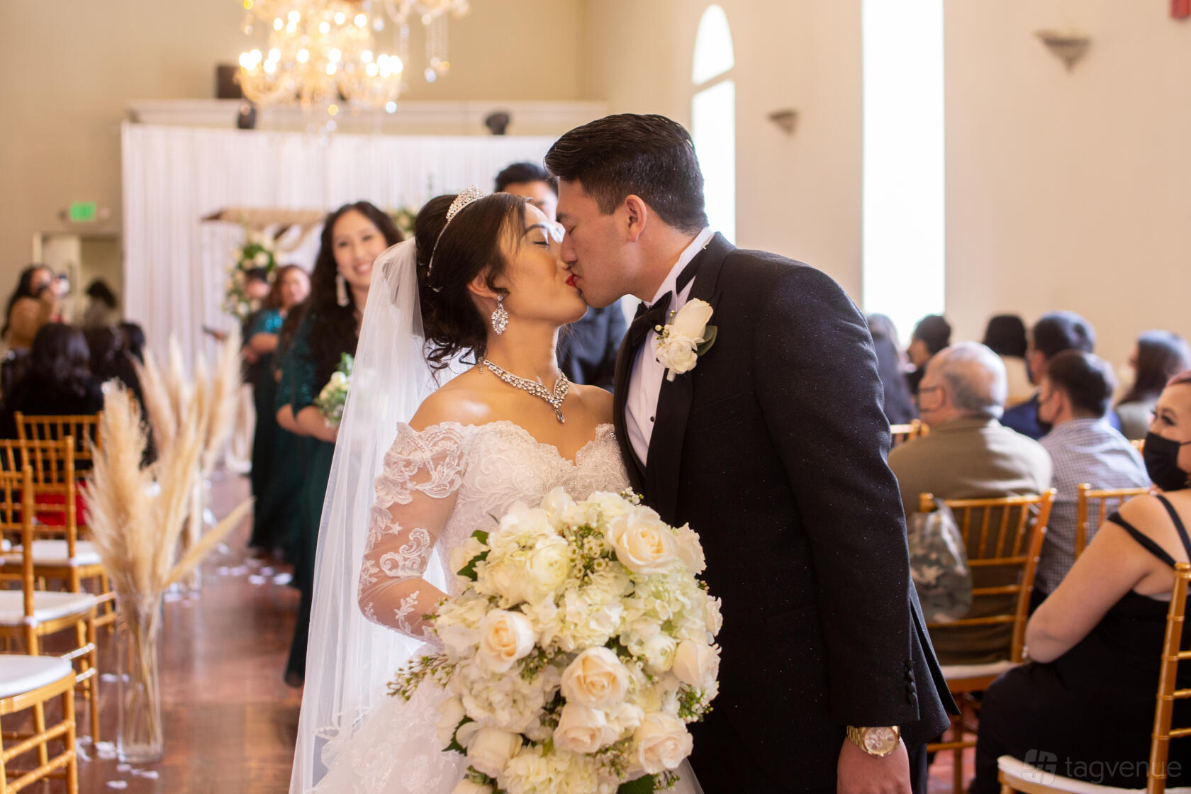 A ballroom with tall arched windows, gold chairs, and a crystal chandelier at Elegant Affair Wedding Venue.