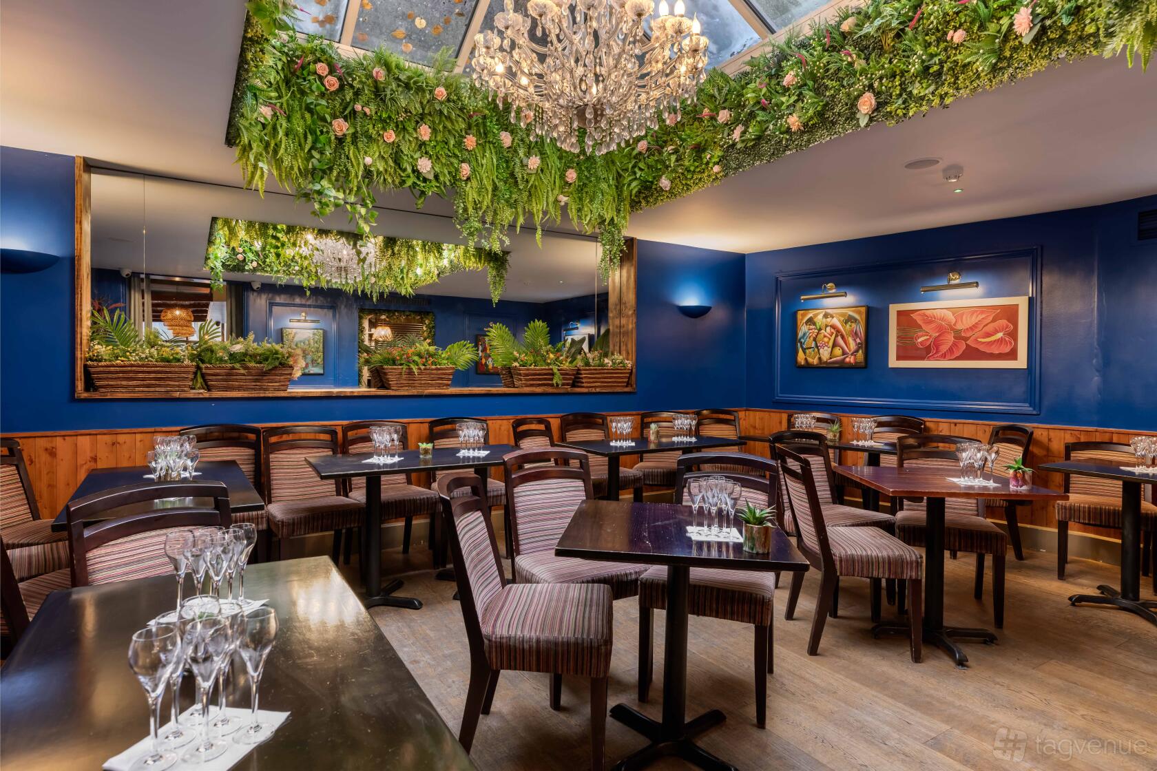 A dining room with a glass ceiling, chandeliers, hanging greenery, blue walls, and wooden tables at Cottons Notting Hill.