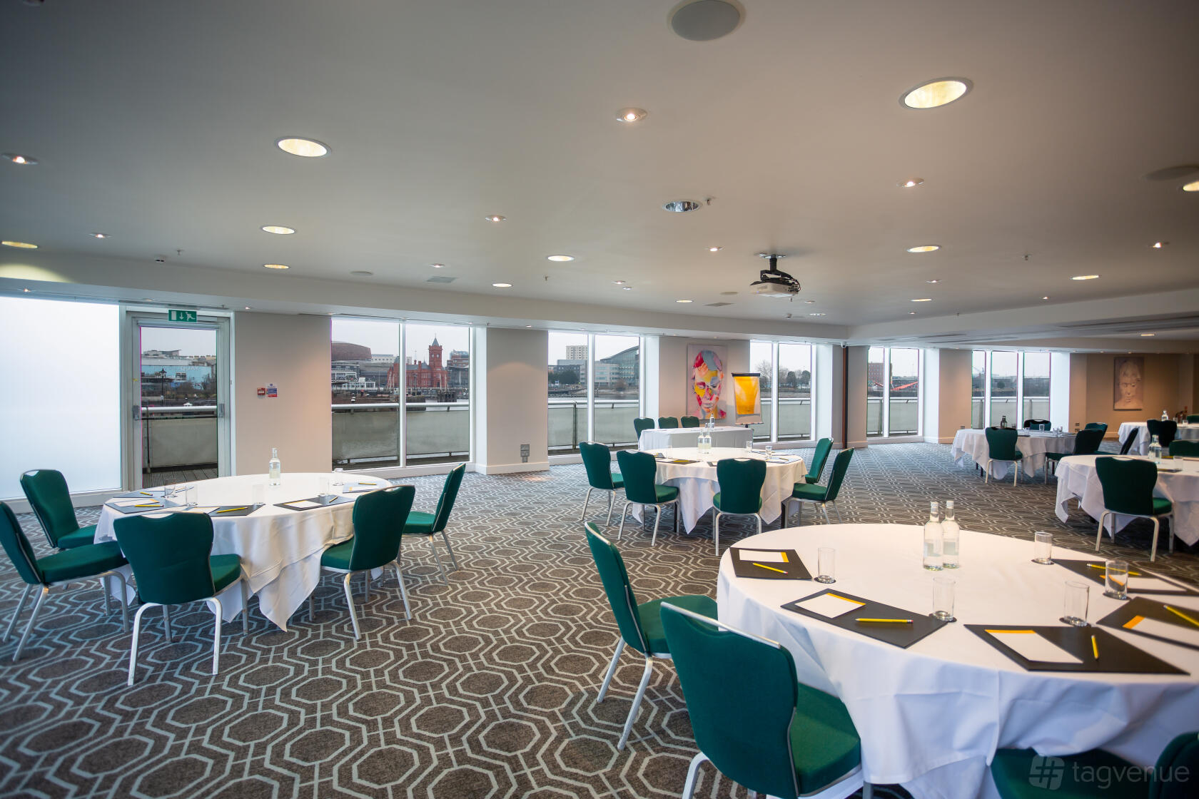 An event space with round tables, green chairs, patterned carpet, and floor-to-ceiling windows at voco St David's Cardiff.