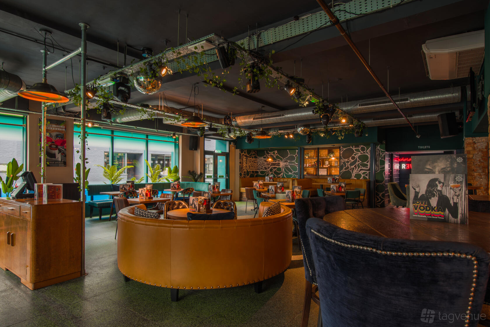 A bar with circular leather seating, exposed ductwork, hanging lights, and large windows at Revolution Southampton.