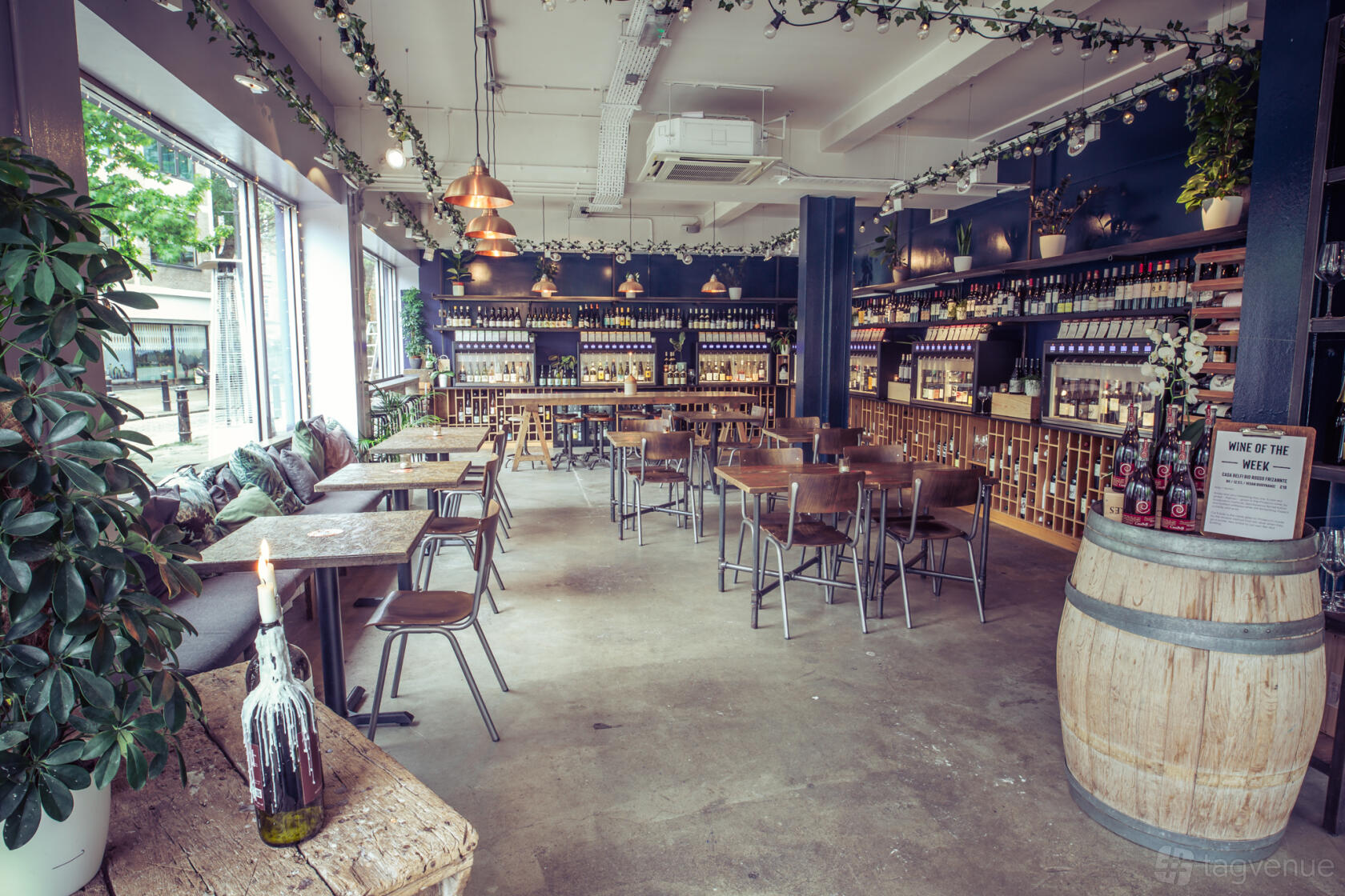 A bar with wooden tables and chairs, wine bottle displays, and hanging pendant lights at Vagabond Fulham.