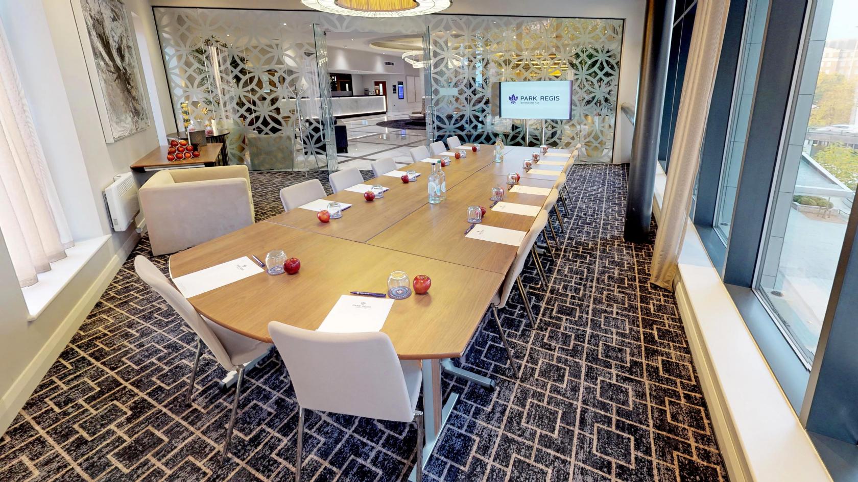 A meeting room with a large boardroom table, cream chairs, patterned carpet, and floor-to-ceiling windows at Park Regis Hotel Birmingham.