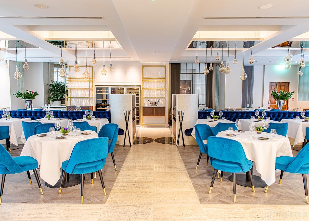 A restaurant dining room with blue velvet chairs, round tables with white linens, and pendant lighting at CORD Restaurant.