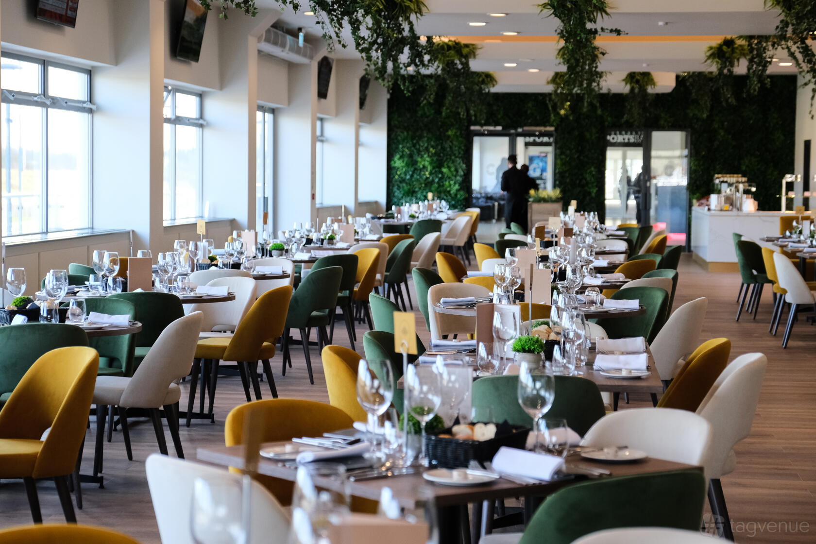 An event venue with floor-to-ceiling windows, greenery accents, and round tables set for dining at Chelmsford City Racecourse.