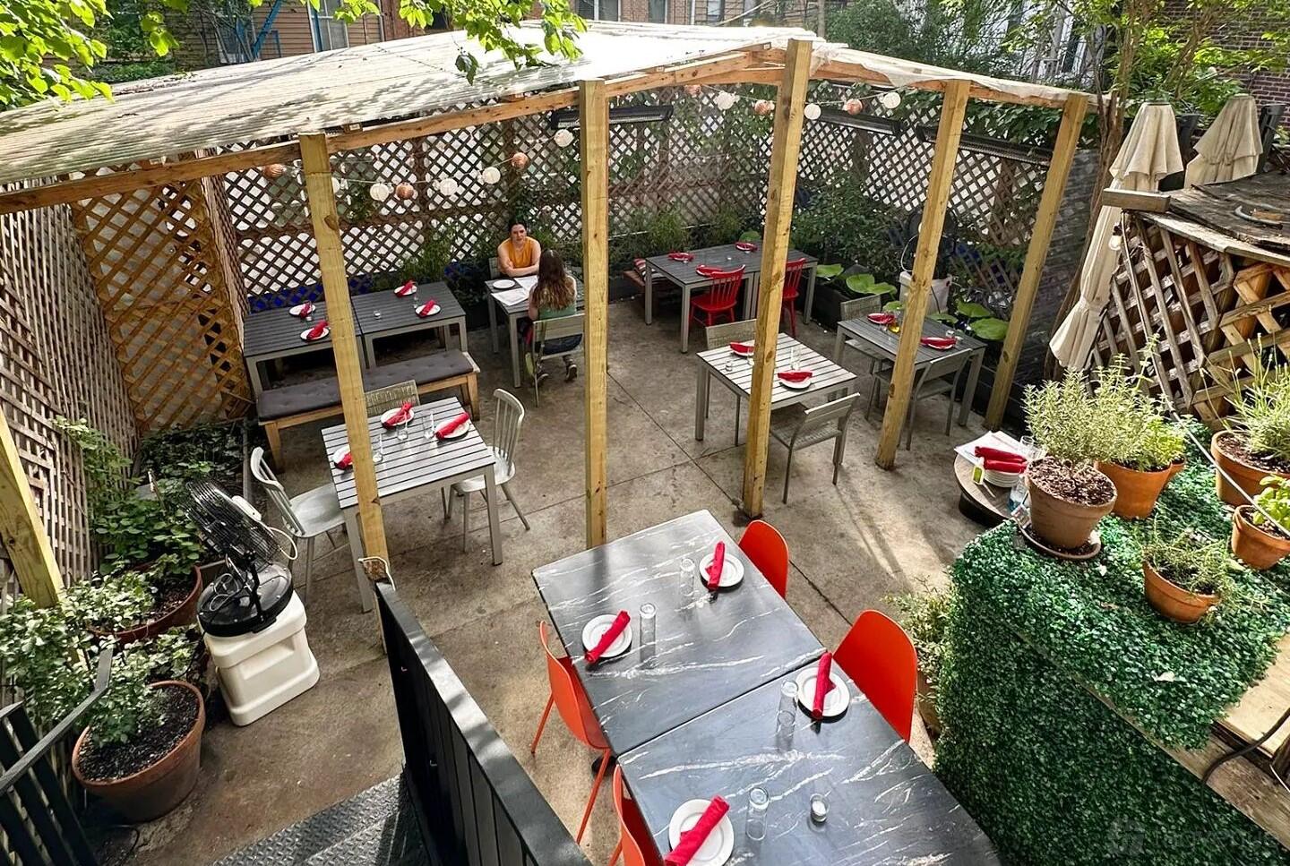 An outdoor restaurant patio with wooden pergola, potted plants, and tables set with red napkins at El Born.