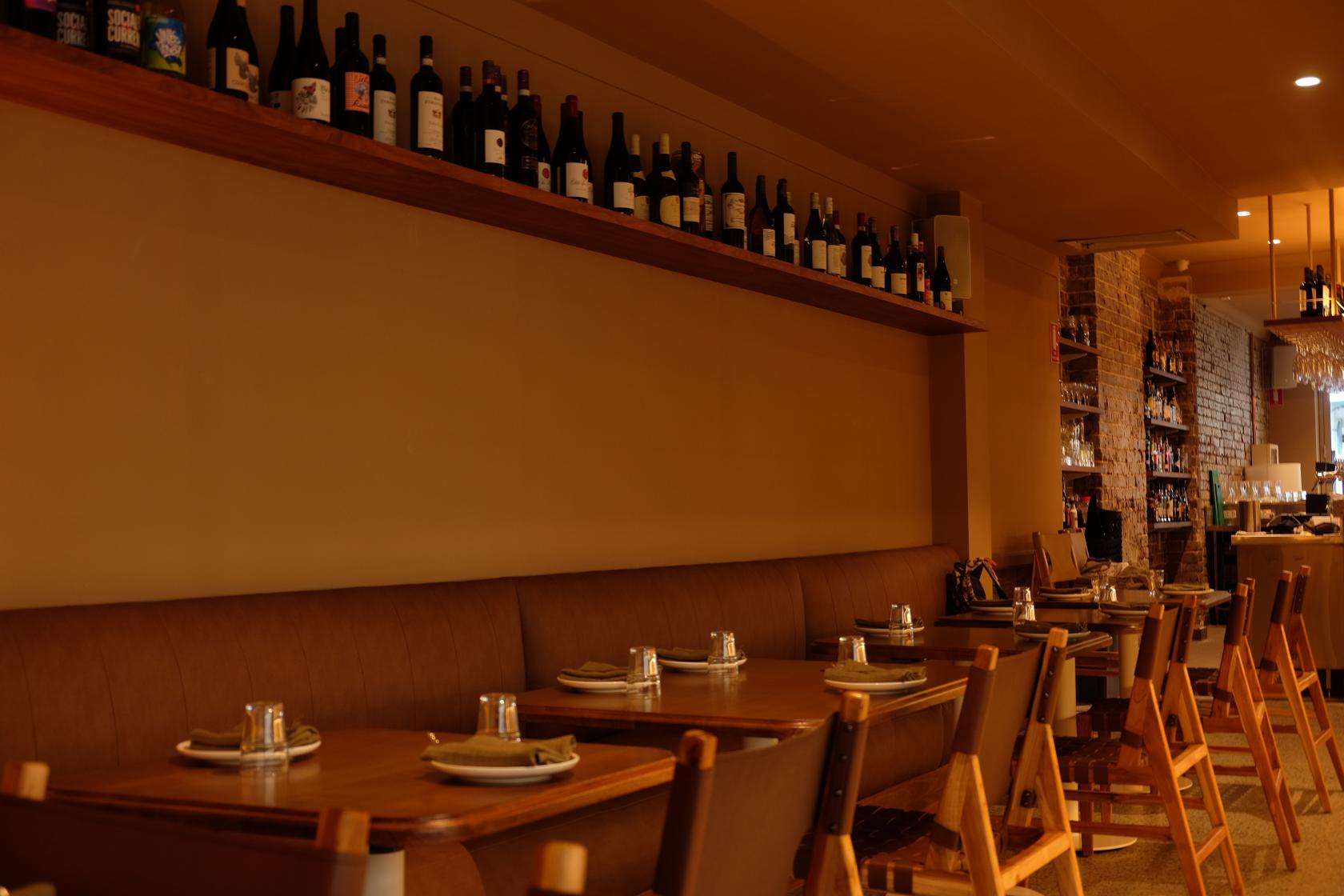 A dining room with wooden tables, cushioned banquette seating, and a display shelf of wine bottles at Yana Coogee.