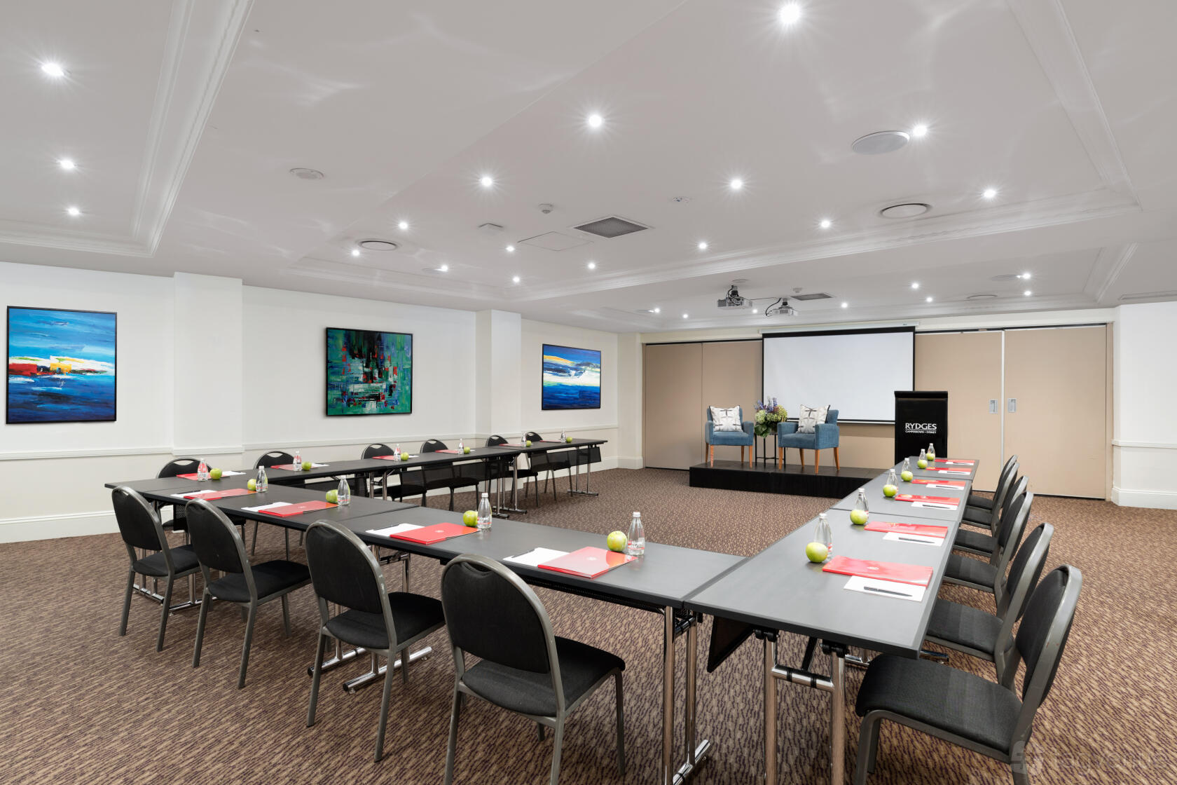 A hotel meeting room with U-shaped tables, black chairs, a projector screen, and modern wall art at Rydges Camperdown.