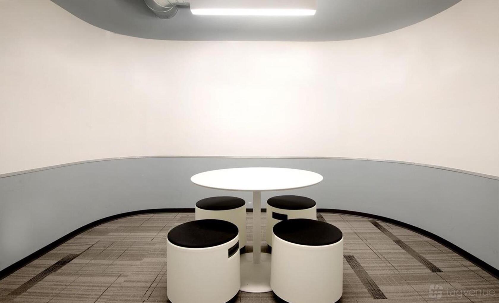A meeting room with a round white table and four black and white stools on patterned carpet at Spark Spaces.