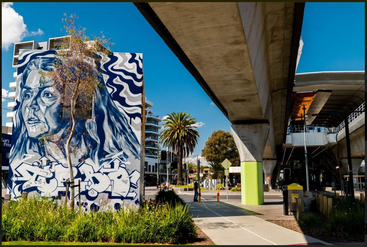 An outdoor area with a large mural, palm trees, and elevated train tracks at Workplex Meeting Centre.