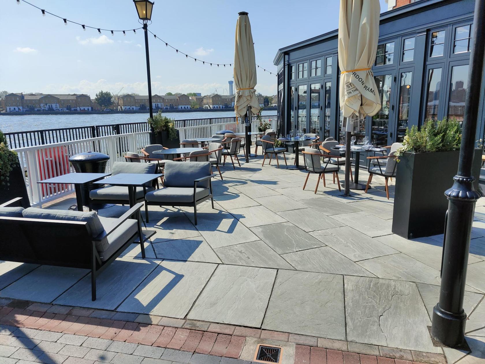 A riverside terrace with outdoor seating, black-framed tables, cushioned chairs, and potted plants at Bread Street Kitchen, Limehouse.