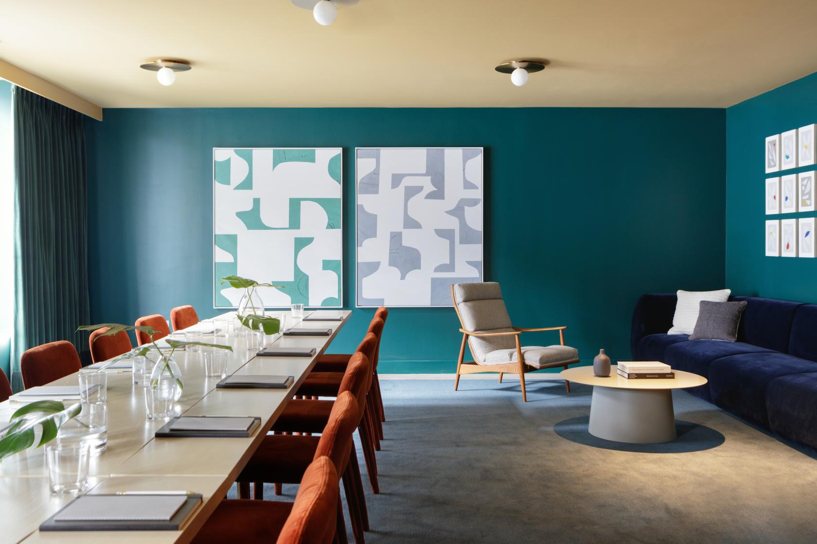 A hotel event room with teal walls, abstract art, a long meeting table with orange chairs, and a lounge area at The Kimpton Sylvan Hotel.
