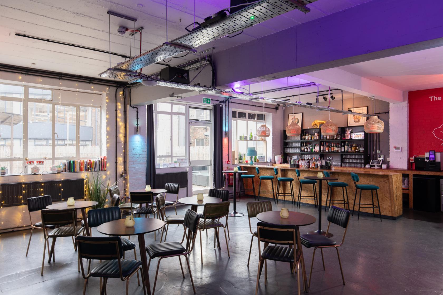 A terrace event space with round tables, black chairs, a wooden bar, and floor-to-ceiling windows at The Drum Labs.