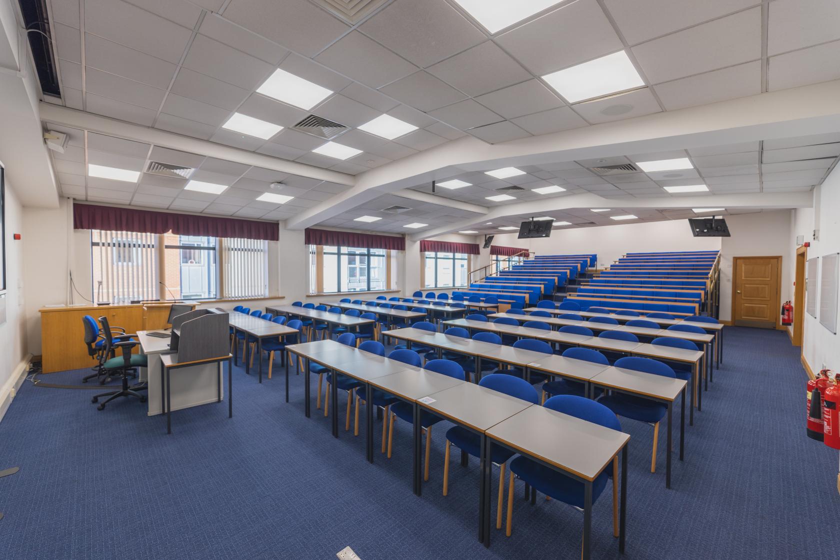 A lecture theatre with tiered rows of blue chairs, long desks, and ceiling-mounted screens at University of Law - Birmingham.