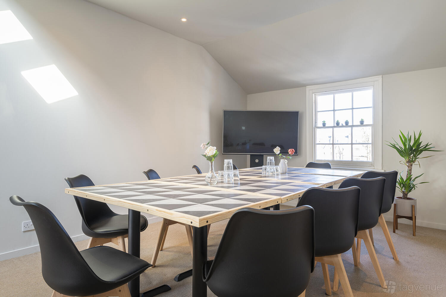 A meeting room with a checkered table, black chairs, large TV screen, and potted plant at Frequency Angel.