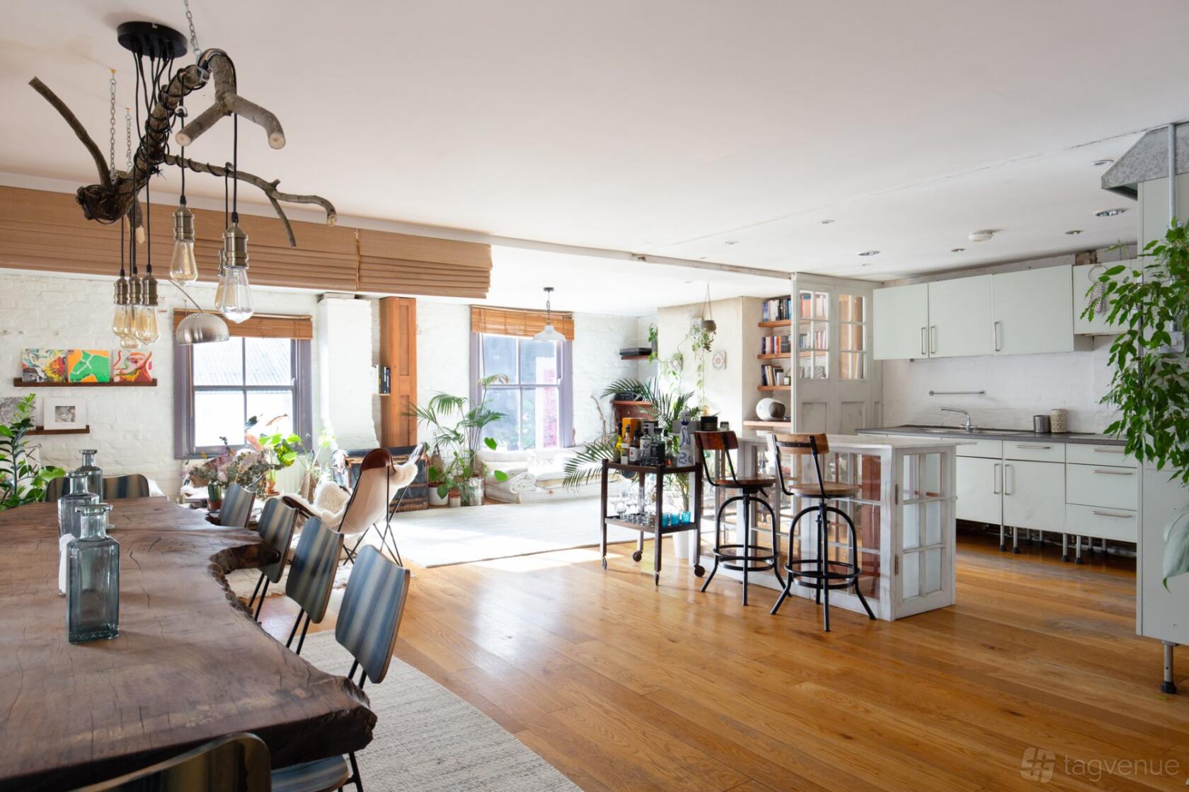 A loft with large windows, wooden floors, a long rustic dining table, kitchen island, and indoor plants at Shoreditch Bohemian townhouse.