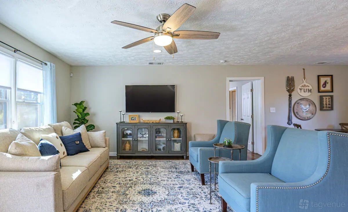 A house living room with blue armchairs, a patterned rug, ceiling fan, large windows, and a wall-mounted TV at Suite Stays Atlanta - Pine Manor, Marietta.