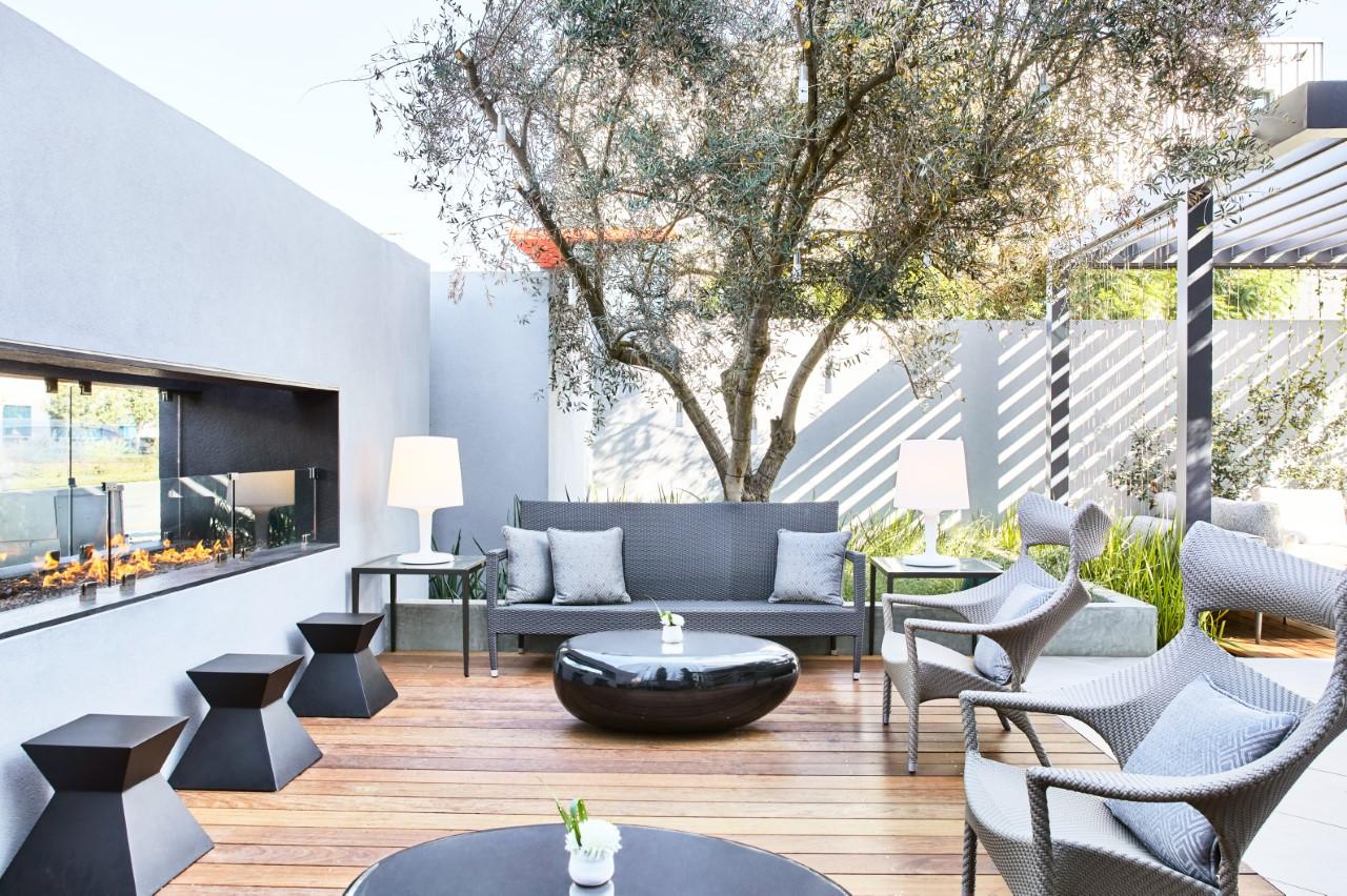 A hotel terrace with wicker lounge seating, a linear fireplace, wood decking, and a tree at AC Hotel Los Angeles South Bay.