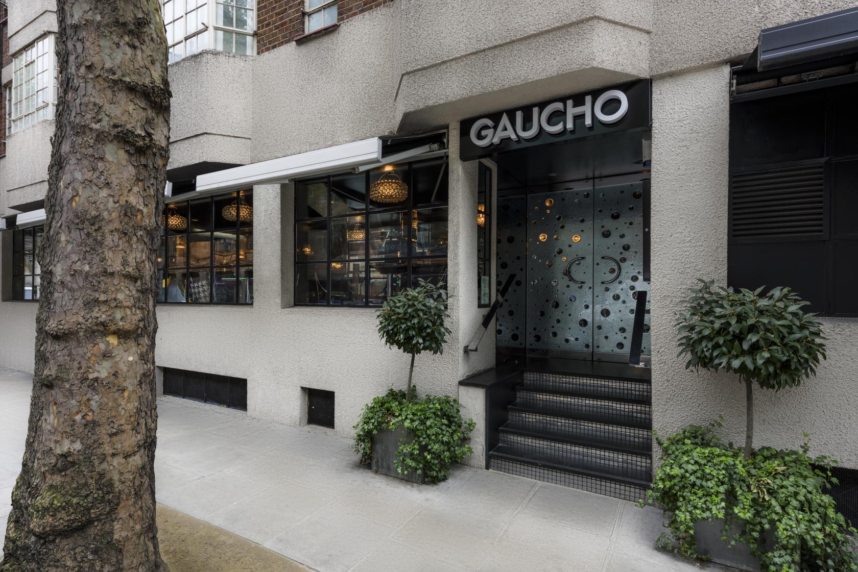 A restaurant entrance with frosted glass doors, black awning, and potted trees at Gaucho Sloane Avenue.