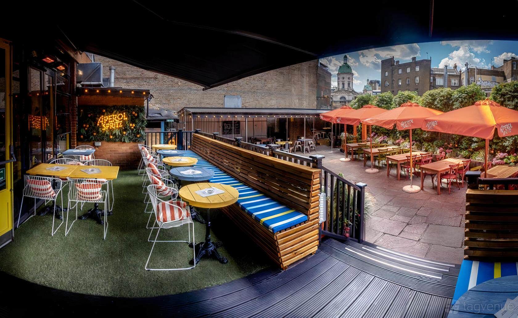 An outdoor pub garden with colorful tables, striped bench seating, and orange umbrellas at The Fence.