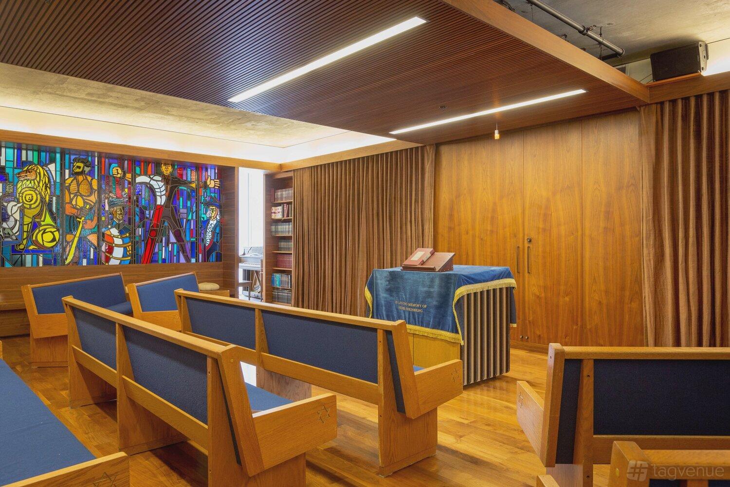 A chapel with wooden pews, blue upholstery, a Torah reading table, and colorful stained glass at Congregation Beth Sholom.