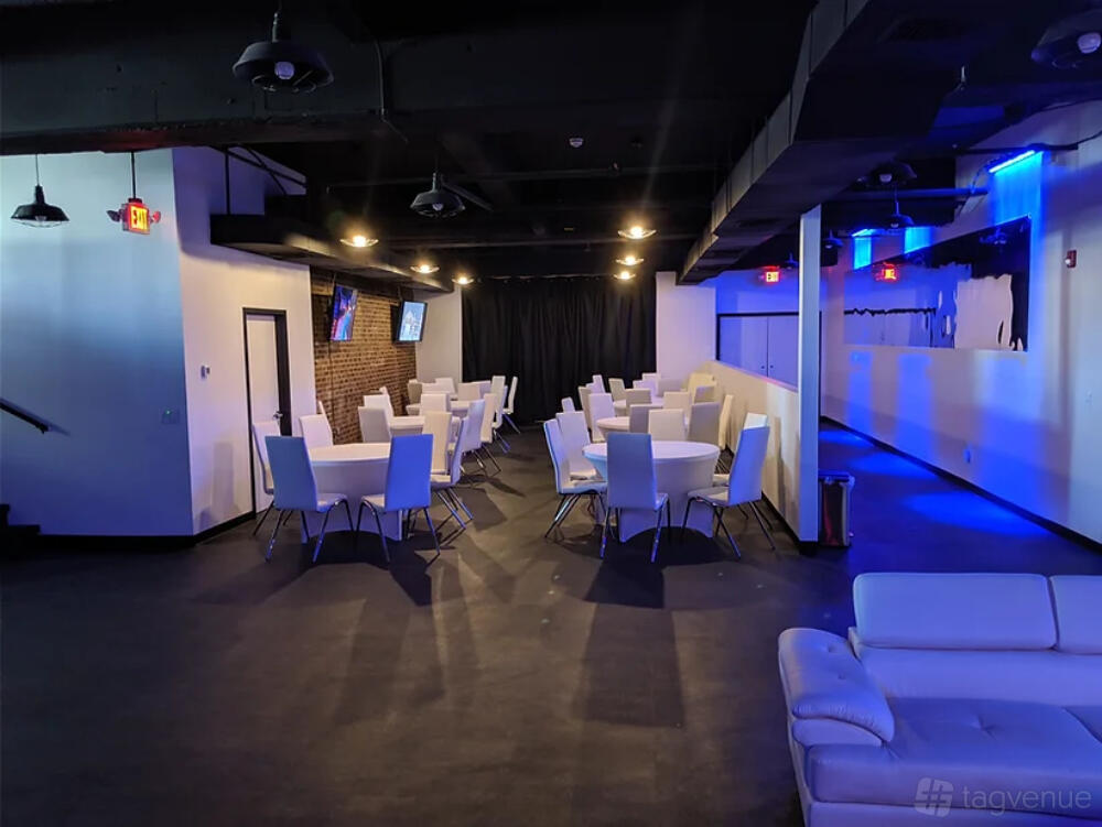 An event venue with white padded chairs, round tables, exposed ceilings, and accent lighting at The Suite Spot.