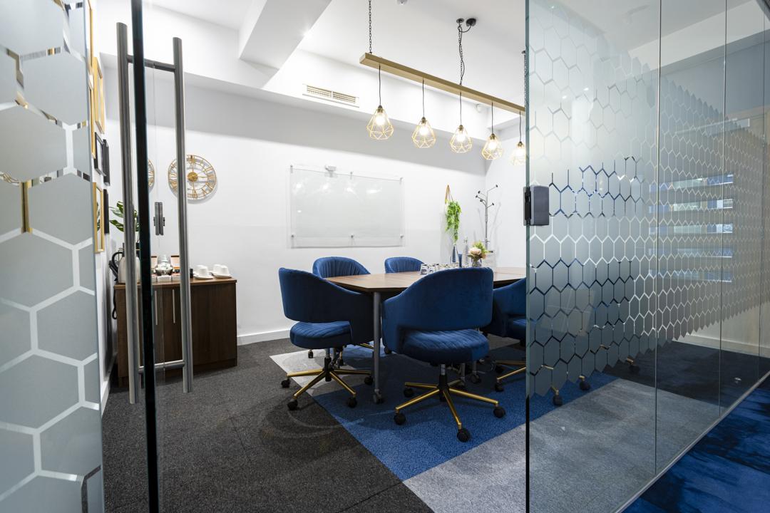 A meeting room with blue cushioned chairs, a wooden table, pendant lighting, and a glass wall at Biz Hub 49 Piccadilly.