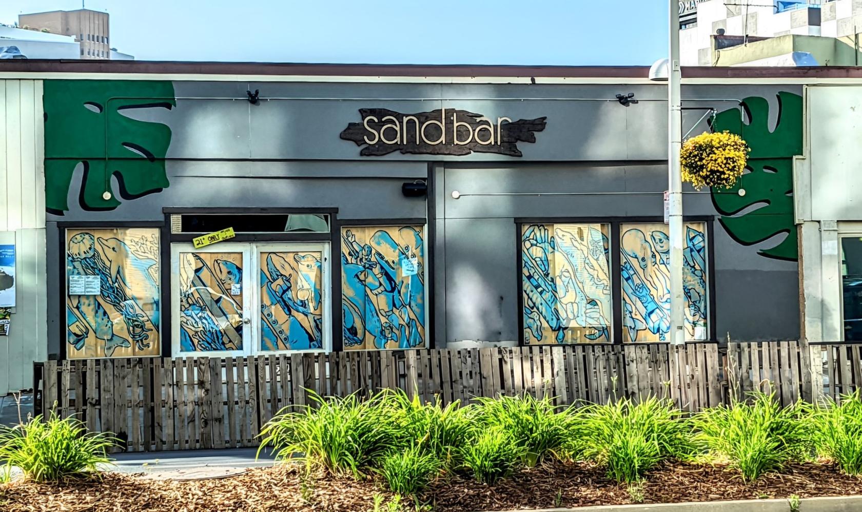A pub with mural-covered windows, tropical leaf accents, and a wood fence at Sandbar.