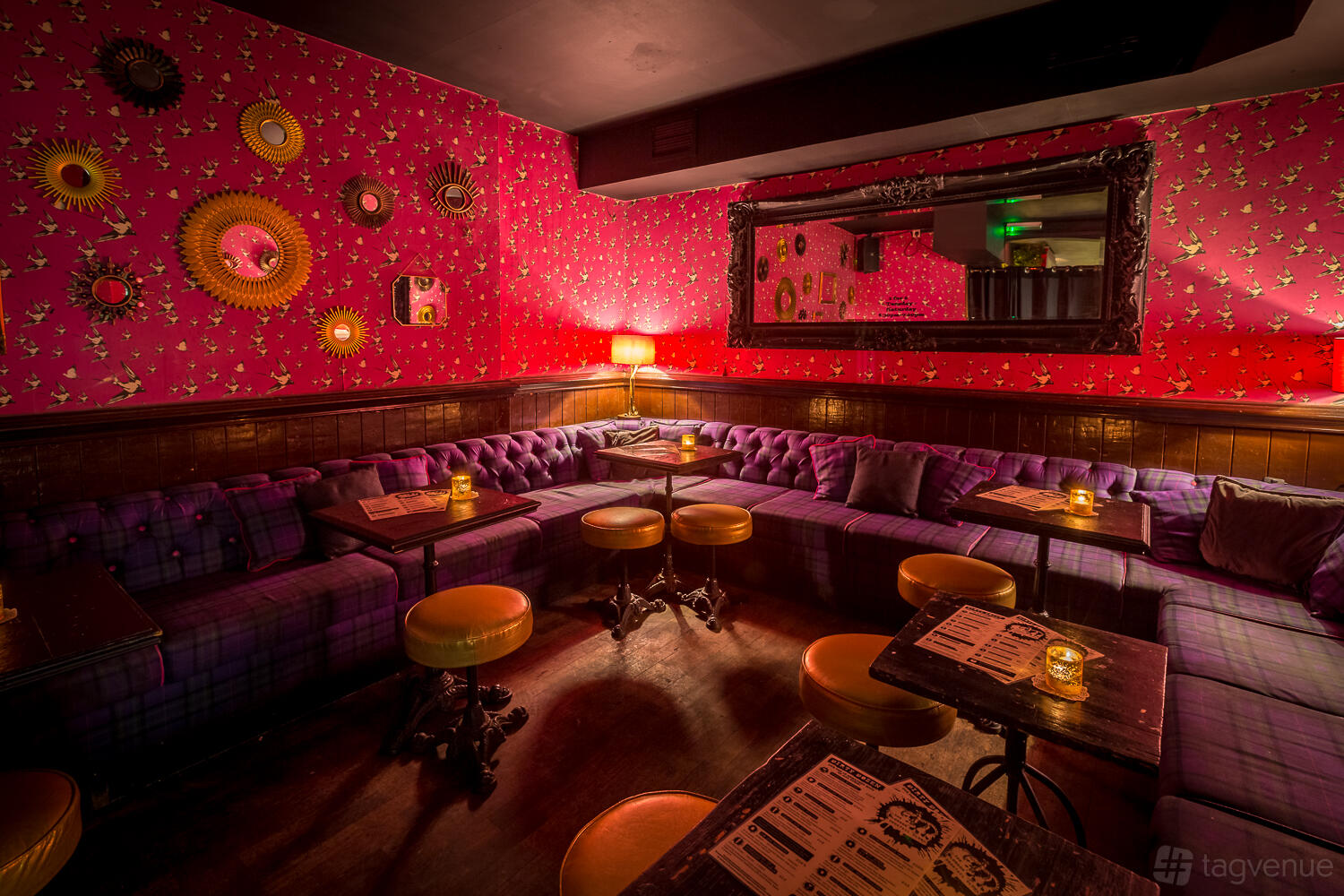 A cocktail bar with patterned pink wallpaper, tufted purple couches, round stools, and dim lighting at The Cocktail Club Goodge Street.