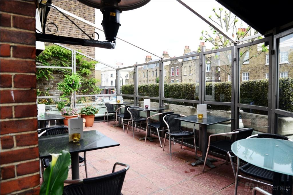 A terrace with glass-topped tables, black chairs, potted plants, and a partial glass canopy at The Chapel Bar.