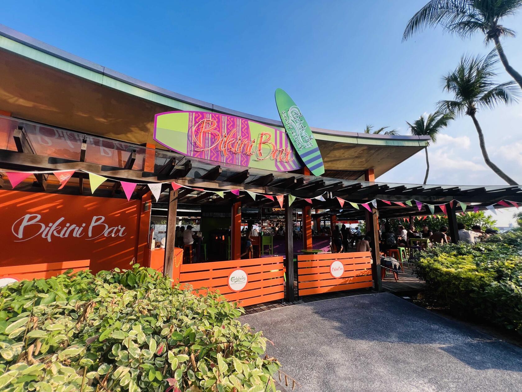 A bar with open-air seating, a colorful sign, surfboard decor, and string flags at Bikini Bar.