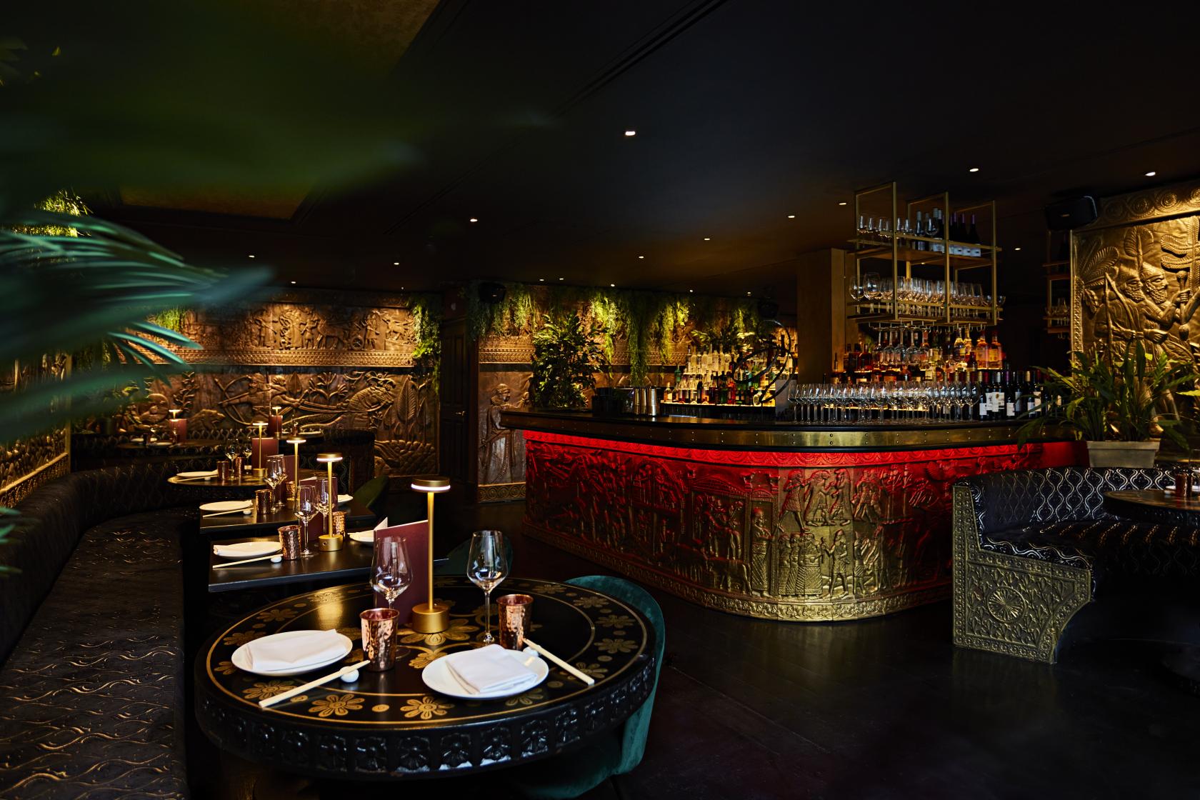 A restaurant with ornate carved walls, red-lit bar, and round tables set with plates and glassware at Gilgamesh London Covent Garden.