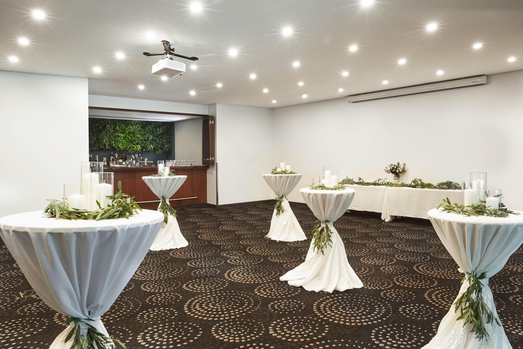A hotel event room with cocktail tables draped in white linens, patterned carpet, and a built-in bar at Crowne Plaza Sydney Airport.