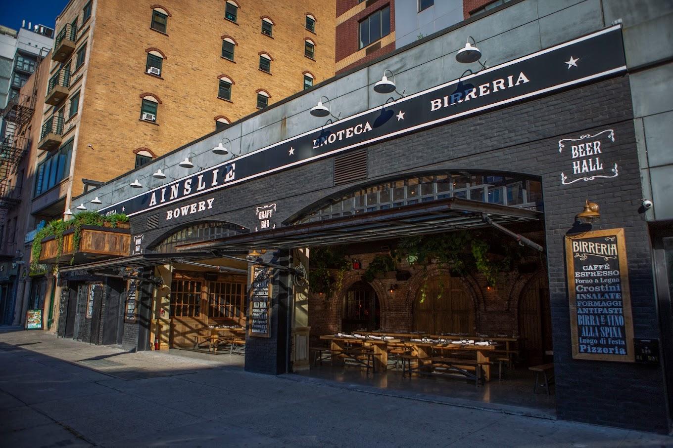 A restaurant with open garage-style doors, exposed brick, and outdoor wooden tables at Ainslie.