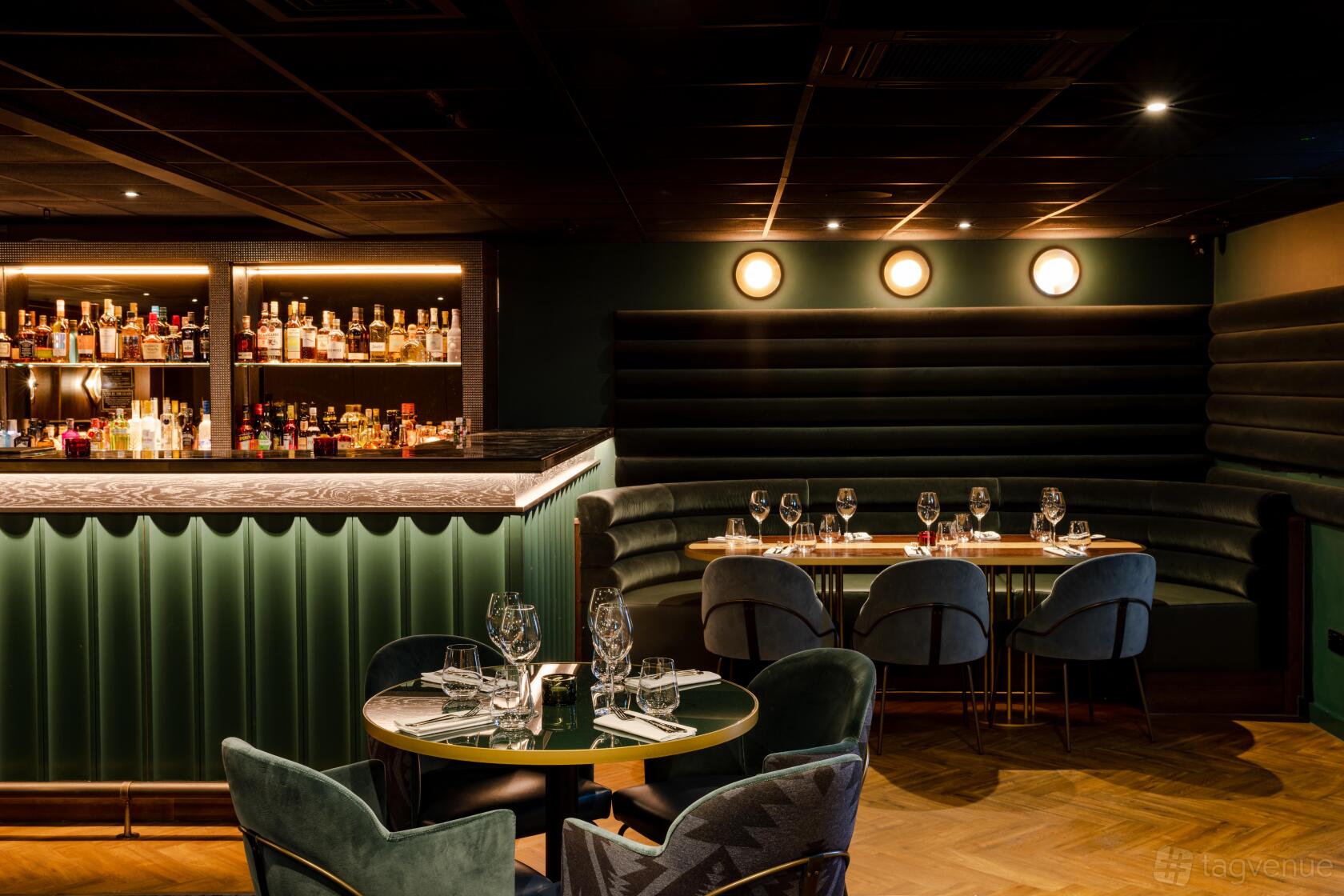 A dining room with velvet banquette seating, dark walls, and a backlit bar display at Gaucho City.