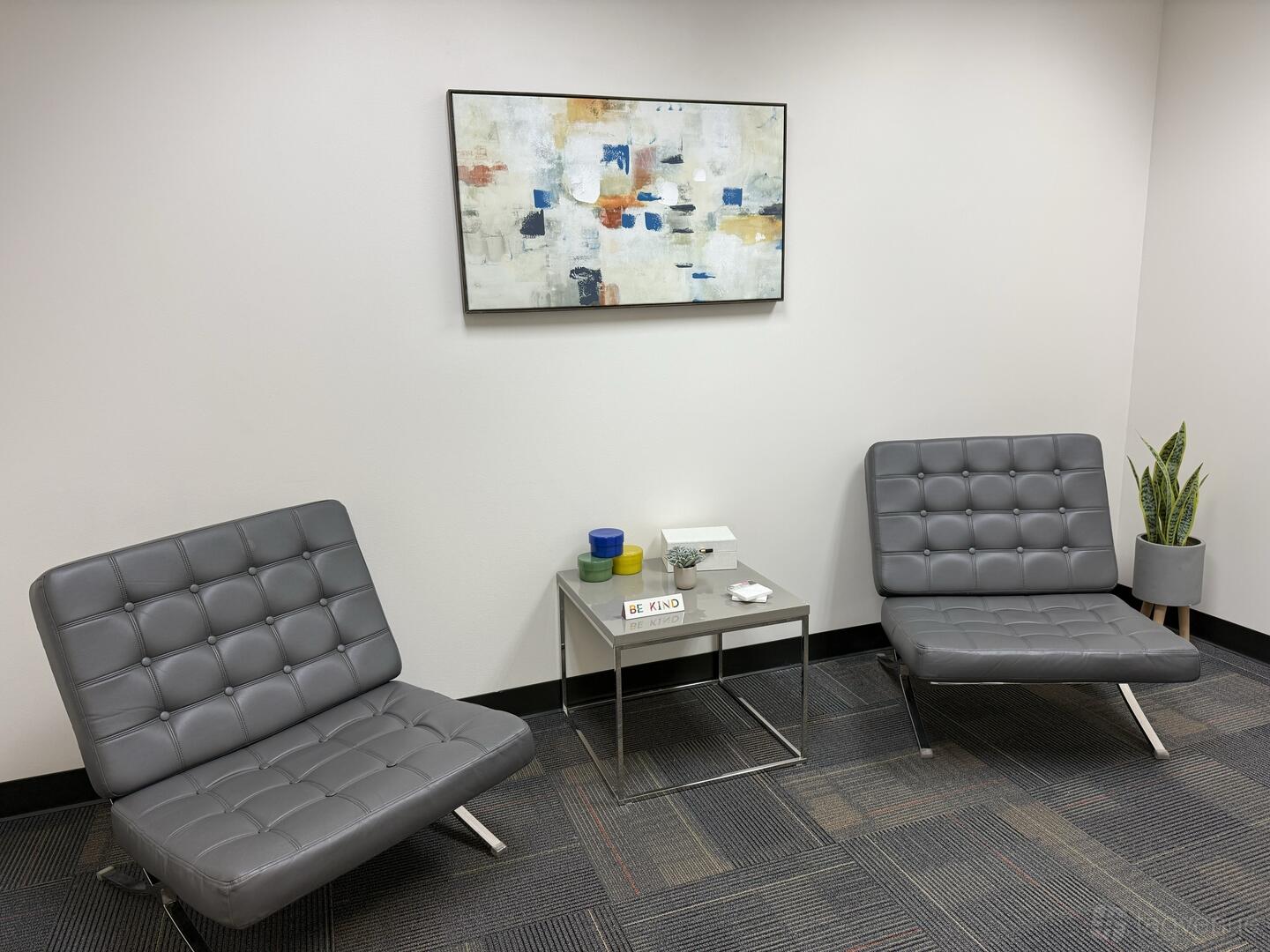A meeting room with gray leather chairs, a small side table, abstract wall art, and a potted plant at Executive Workspace Park Ventura.
