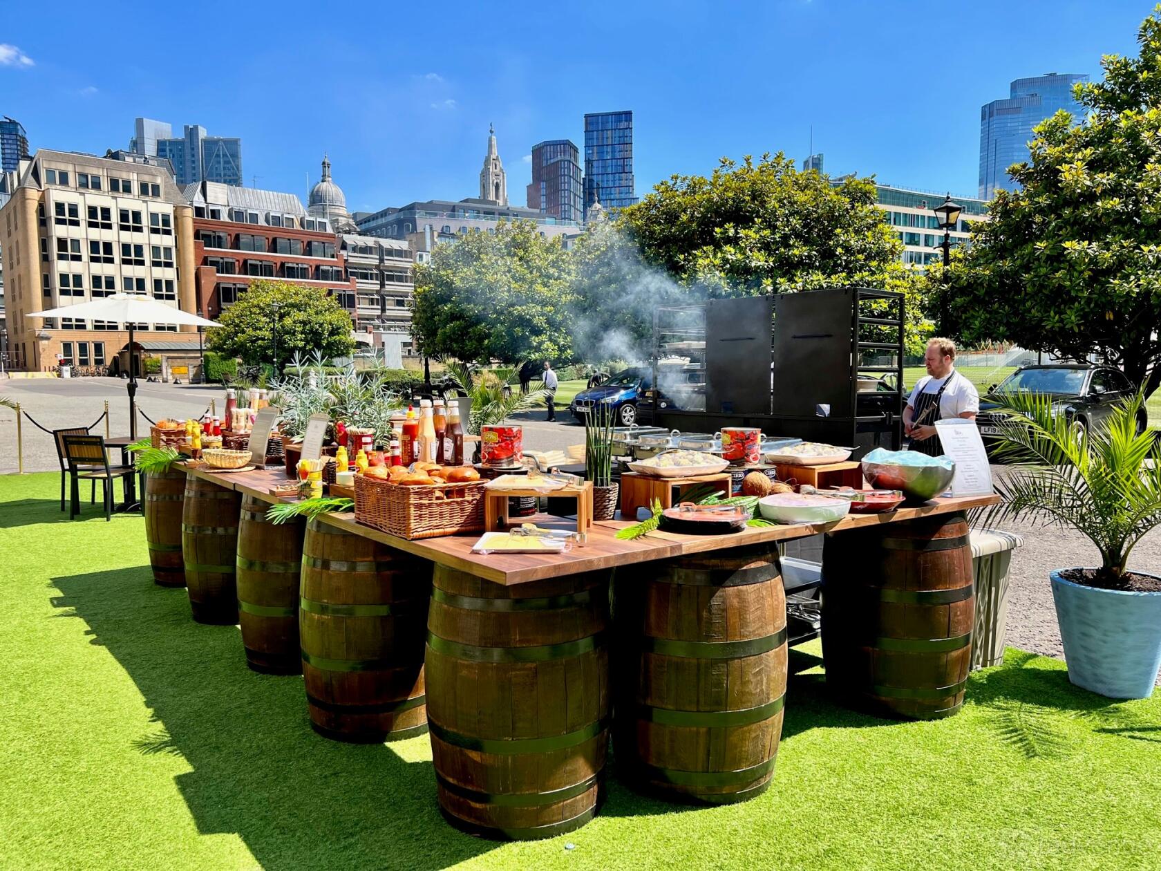 An outdoor event venue with wooden barrel tables, a buffet spread, and city buildings in the background at The HAC.