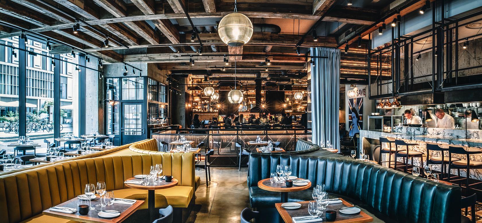 A restaurant with exposed wooden beams, round tables set with glassware, and leather banquette seating at Bread Street Kitchen, The City.