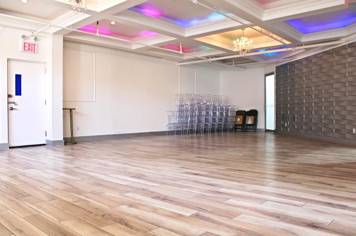 An event venue with wood flooring, stacked clear chairs, and a coffered ceiling with colorful accent lighting at Suite 989 Events.