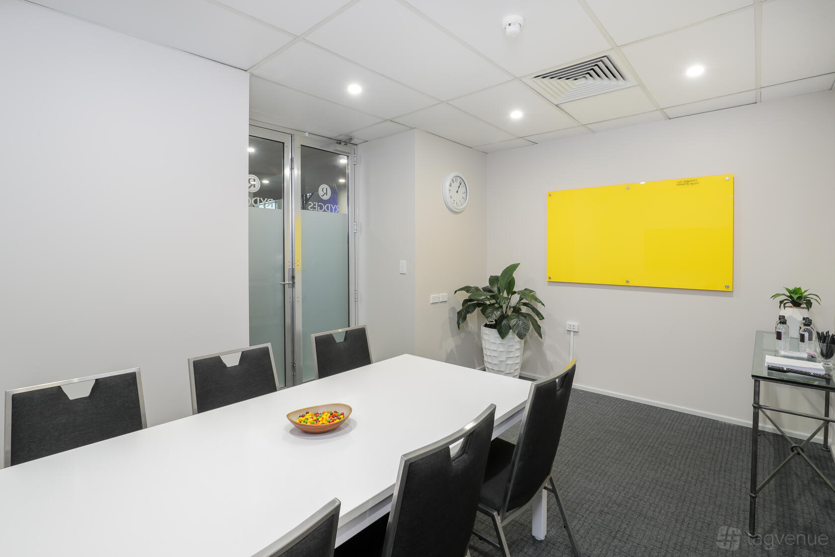 A meeting room with a whiteboard, conference table, black chairs, potted plant, and clock at Rydges Parramatta.
