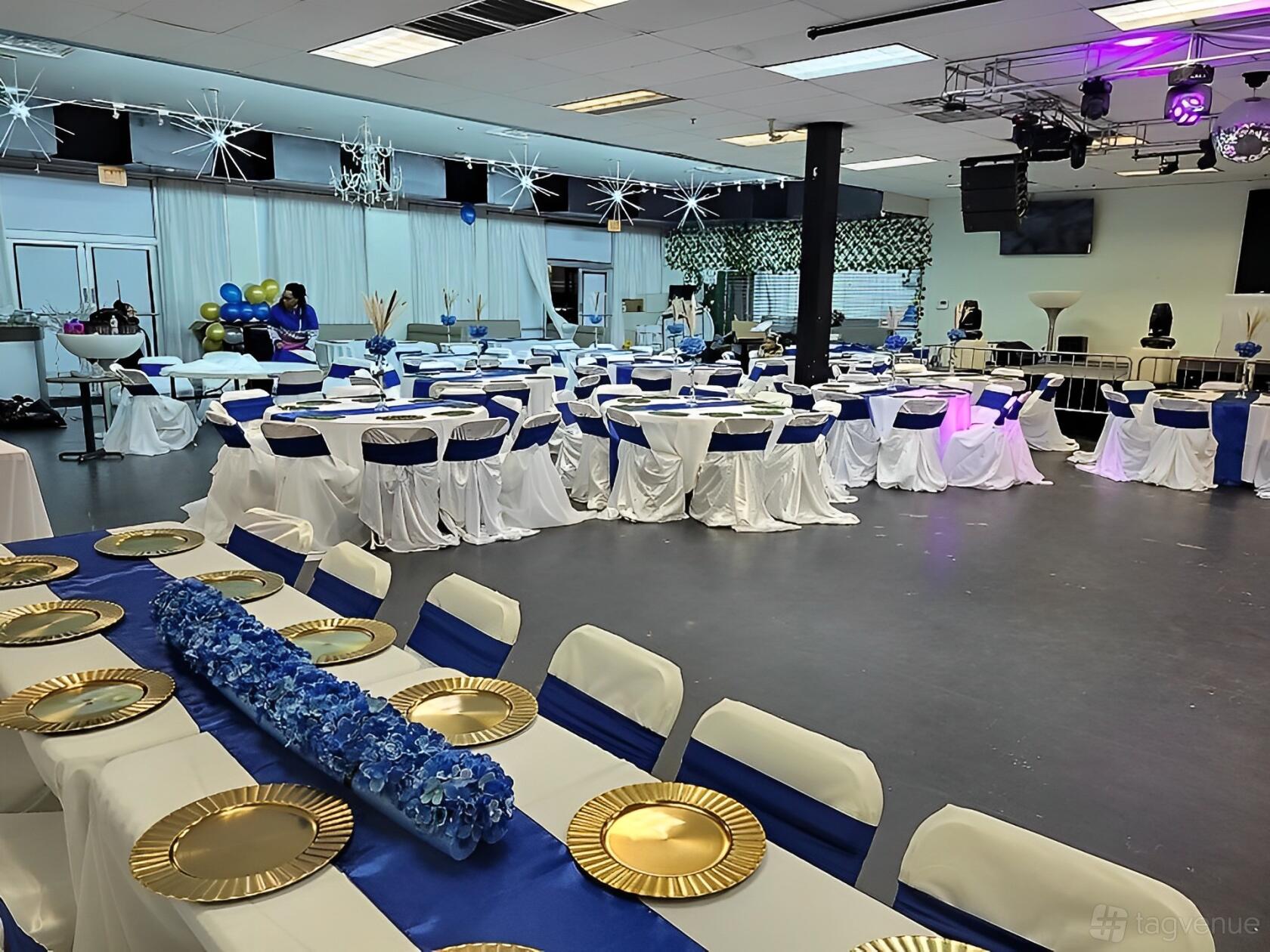An event venue with round tables draped in white linens and blue runners, gold charger plates, and stage lighting at Punto Zero Event Center.