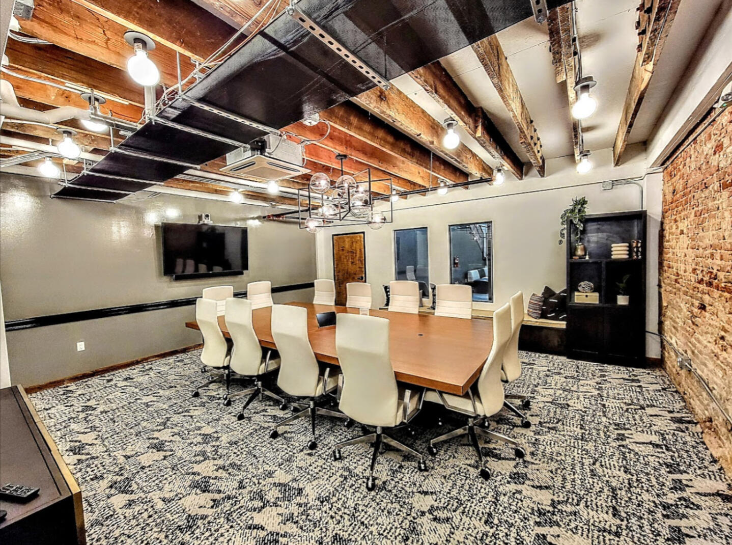 A boardroom with a long wooden table, white leather chairs, exposed brick wall, and ceiling beams at 25N Coworking Waco.