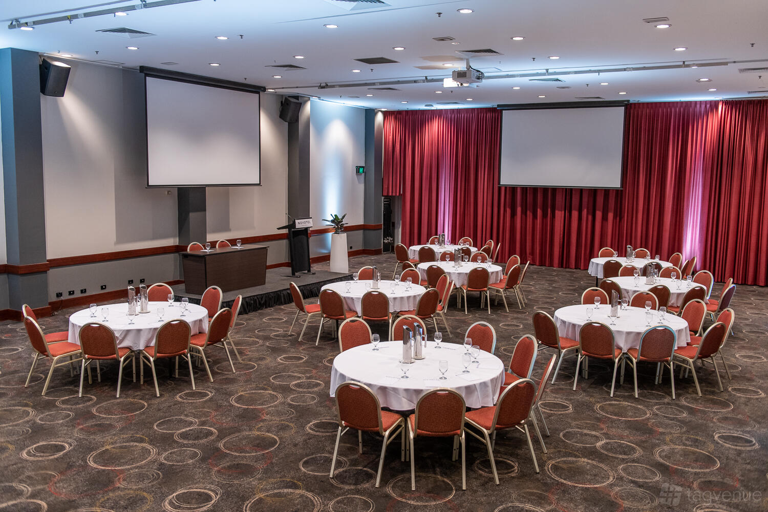 A hotel ballroom with round tables, red chairs, ceiling-mounted projectors, and red curtains at Novotel Perth Langley.