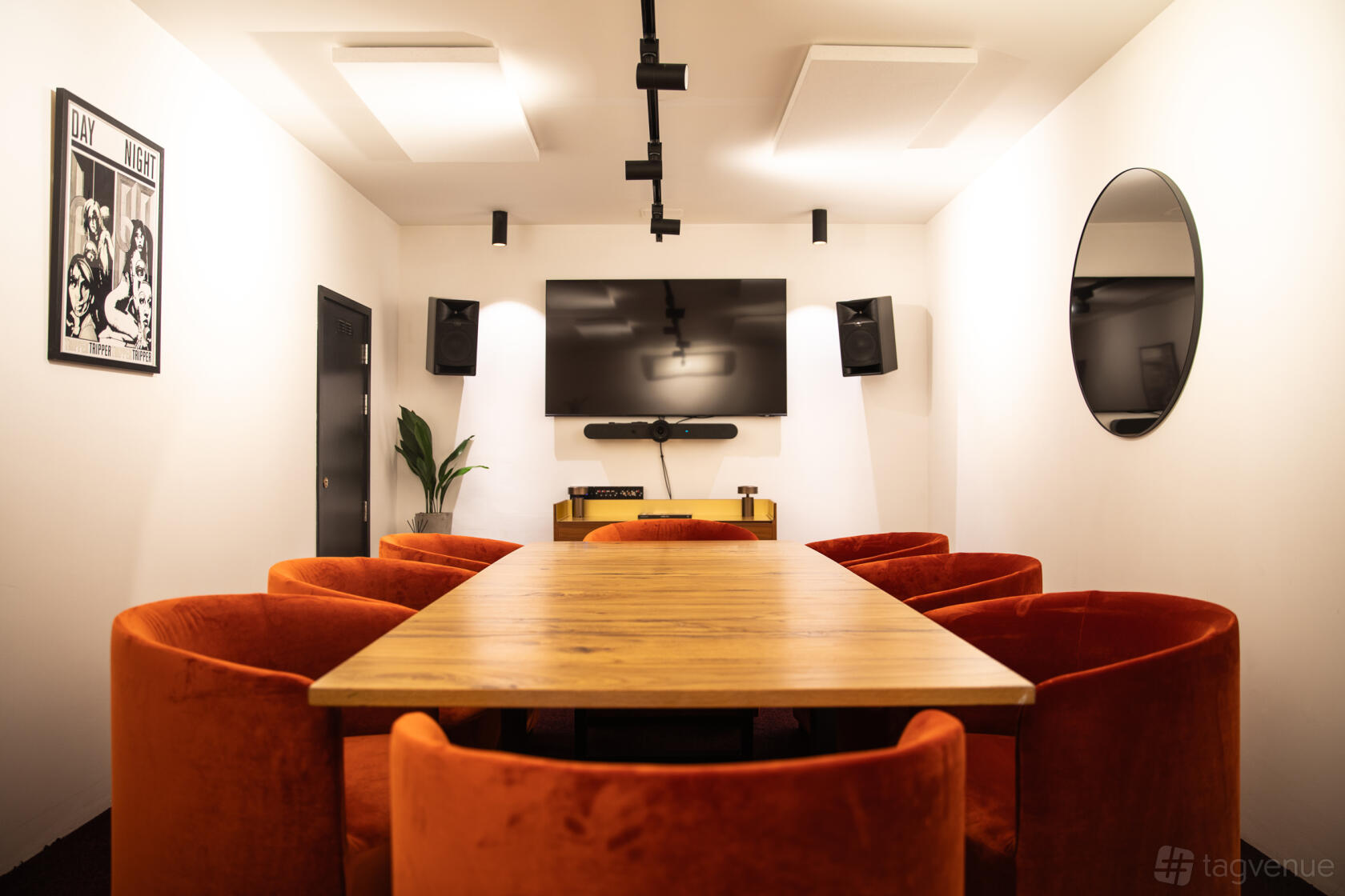 A meeting room with a long wooden table, orange velvet chairs, wall-mounted screen, and circular mirror at The Ministry.