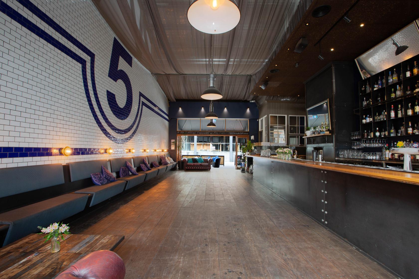 A bar with a long wooden counter, exposed ceiling, white tiled wall with a large number 5, and lounge seating at Plus 5.