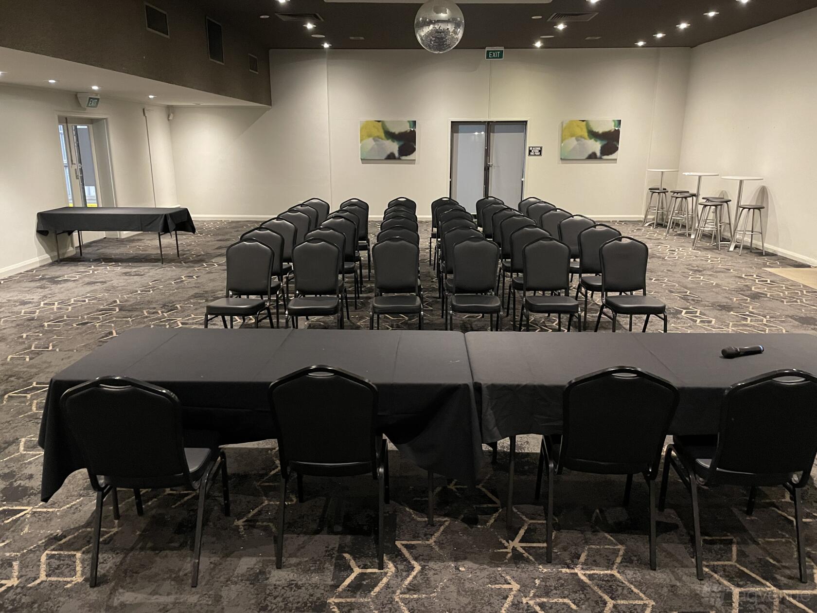 A pub function room with rows of black chairs, tables with black linens, patterned carpet, and abstract wall art at Sandbelt Club Hotel.