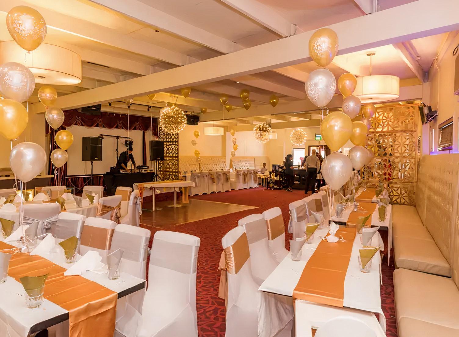 A restaurant with long banquet tables covered in gold runners, white chair covers, and balloons at Tandoori Flames Restaurant.