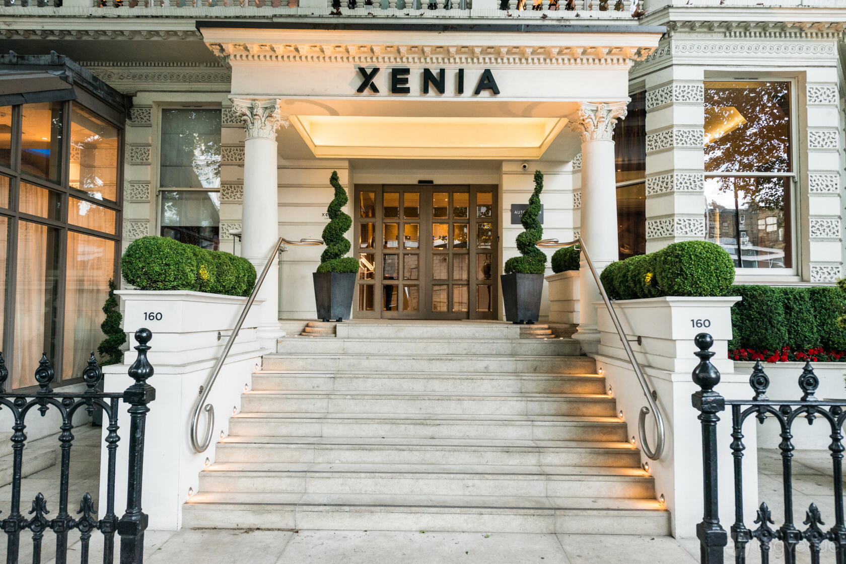 A hotel entrance with wide stone steps, topiary plants, and large glass windows at Hotel Xenia.