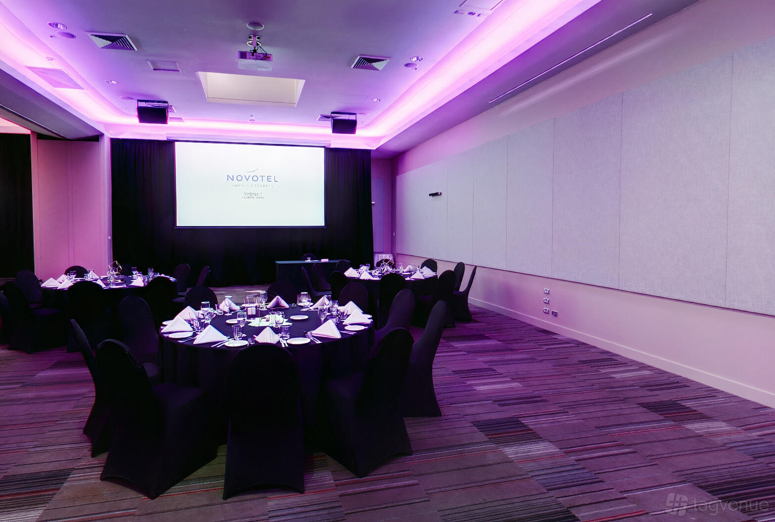 A meeting room with round tables draped in black linens, purple uplighting, and a projector screen at Novotel Sydney Olympic Park Hotel.