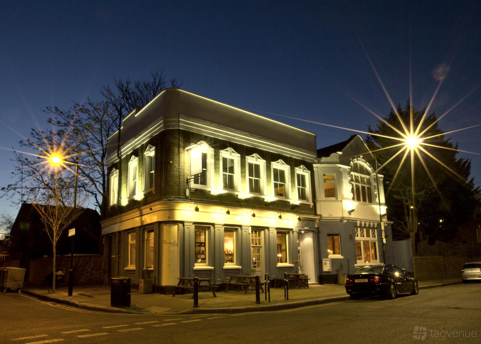 An illuminated pub with large windows, outdoor benches, and street lighting at The Crooked Well.